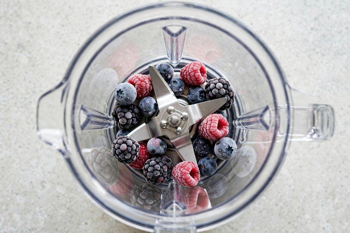 Stainless blender blades seen from above inside a clear pitcher, frozen berries resting on the blades